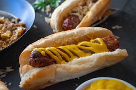 A grilled sausage in a bun topped with yellow mustard, alongside another sausage with sauerkraut in the background. A pan with fried onions and a dish with more mustard are present, suggesting a meal preparation setting.