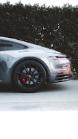 A sleek silver sports car is parked on a pavement next to green hedges. The focus is on the car's rear section, highlighting the black alloy wheels with red brake calipers and the smooth curves of the vehicle's body. The design and details suggest a modern, luxurious automotive aesthetic.