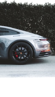 A sleek silver sports car is parked on a pavement next to green hedges. The focus is on the car's rear section, highlighting the black alloy wheels with red brake calipers and the smooth curves of the vehicle's body. The design and details suggest a modern, luxurious automotive aesthetic.