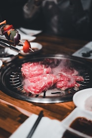 Slices of marbled meat are cooking on a tabletop grill, with steam rising from the grill. A skewer with colorful vegetables, including tomatoes and bell peppers, is held above the grill. The table is laid out with various sauces and napkins, suggesting a dining setup.