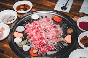 A variety of thinly sliced marinated meats and vegetables arranged on a grill. Surrounding the grill are small side dishes containing pickled vegetables and sauces.