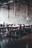 A modern dining area with industrial-style decor. The space features long wooden tables and black metal chairs, all arranged neatly in rows. Metal ceiling fixtures add an industrial touch, and the concrete walls contribute to the minimalist aesthetic. The room is lit by natural light, enhancing the warm tones of the wood.