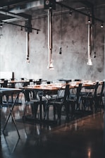 A modern dining area with industrial-style decor. The space features long wooden tables and black metal chairs, all arranged neatly in rows. Metal ceiling fixtures add an industrial touch, and the concrete walls contribute to the minimalist aesthetic. The room is lit by natural light, enhancing the warm tones of the wood.