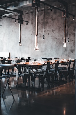A modern dining area with industrial-style decor. The space features long wooden tables and black metal chairs, all arranged neatly in rows. Metal ceiling fixtures add an industrial touch, and the concrete walls contribute to the minimalist aesthetic. The room is lit by natural light, enhancing the warm tones of the wood.