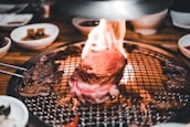 A thick piece of steak is sizzling on a grill with flames around it, surrounded by various side dishes in small bowls. The setting suggests a dining experience focused on grilling meat with an open flame.