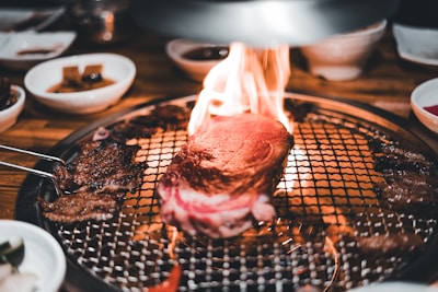A thick piece of steak is sizzling on a grill with flames around it, surrounded by various side dishes in small bowls. The setting suggests a dining experience focused on grilling meat with an open flame.