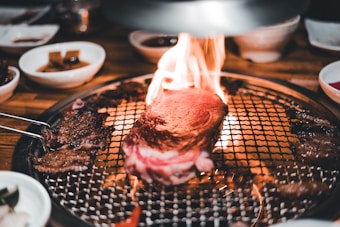 A thick piece of steak is sizzling on a grill with flames around it, surrounded by various side dishes in small bowls. The setting suggests a dining experience focused on grilling meat with an open flame.