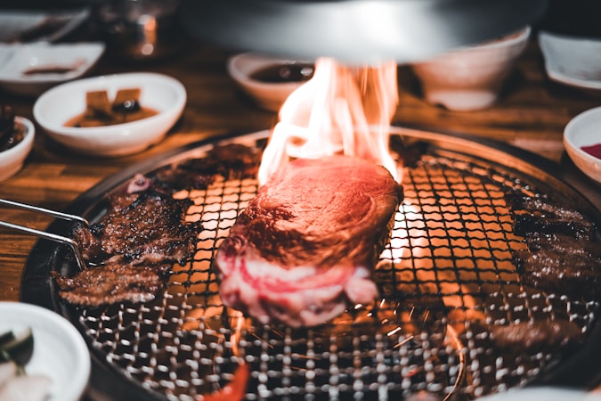 A thick piece of steak is sizzling on a grill with flames around it, surrounded by various side dishes in small bowls. The setting suggests a dining experience focused on grilling meat with an open flame.