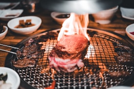 A thick piece of steak is sizzling on a grill with flames around it, surrounded by various side dishes in small bowls. The setting suggests a dining experience focused on grilling meat with an open flame.