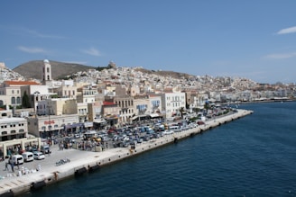 A coastal town with densely packed buildings along the waterfront, a mix of modern and traditional architecture. Cars are parked along the waterfront promenade, and a few people are seen walking. The sea is calm with a deep blue color, and the hill in the background is dotted with houses and a church bell tower.