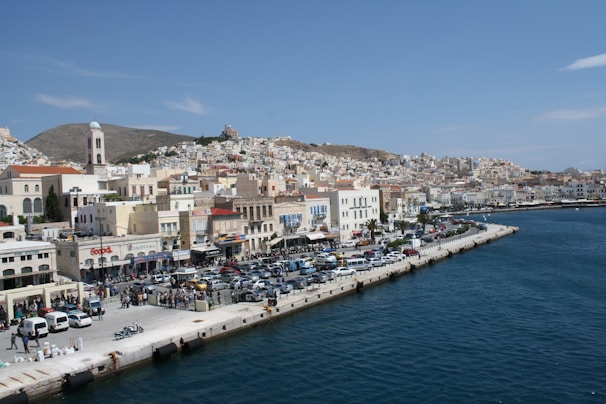 A coastal town with densely packed buildings along the waterfront, a mix of modern and traditional architecture. Cars are parked along the waterfront promenade, and a few people are seen walking. The sea is calm with a deep blue color, and the hill in the background is dotted with houses and a church bell tower.