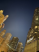 A nighttime cityscape featuring illuminated skyscrapers with various architectural designs against a dark blue sky. Prominent among the buildings is one displaying the word 'TRUMP'. The lights from the buildings create a glowing effect against the night.