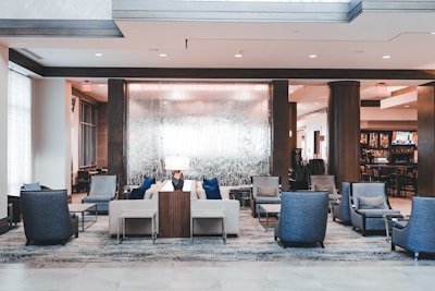 The image shows a modern and elegant hotel lobby with a luxurious seating area. Several cushioned chairs and sofas are arranged neatly around a low coffee table, upon which a decorative lamp is positioned. The flooring features a patterned carpet, while the walls and ceiling are adorned with wooden accents and contemporary light fixtures. In the background, there is a semi-transparent decorative partition that separates the seating area from a bar or restaurant space.