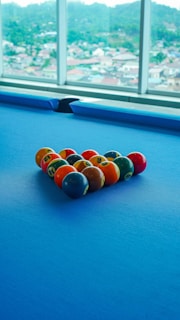 A set of colorful billiard balls arranged in a triangle on a blue pool table. Large windows in the background reveal a blurred view of a cityscape with hills in the distance.