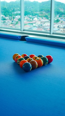 A set of colorful billiard balls arranged in a triangle on a blue pool table. Large windows in the background reveal a blurred view of a cityscape with hills in the distance.