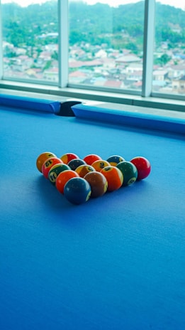 A set of colorful billiard balls arranged in a triangle on a blue pool table. Large windows in the background reveal a blurred view of a cityscape with hills in the distance.