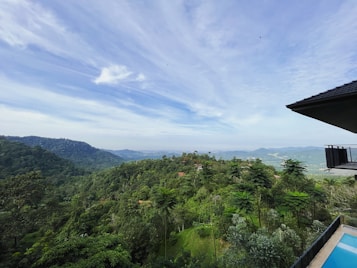 A vast, lush green landscape with dense forest covering rolling hills. The sky is bright and expansive with wispy clouds. In the foreground, a modern house balcony with a swimming pool is seen, offering a panoramic view of the surroundings.