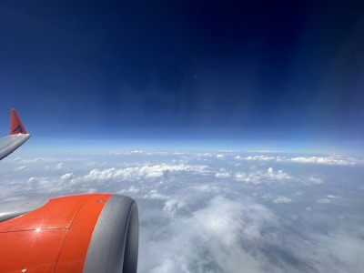 An airplane wing and engine are visible against a backdrop of a clear blue sky and scattered white clouds.