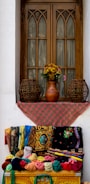 A wooden window with arched design elements is adorned with a red patterned cloth. Below the window, an array of colorful fabrics and textiles, featuring intricate patterns, is neatly displayed. Two woven wicker baskets flank a vase with flowers on the windowsill, adding charm to the setting.