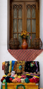 A wooden window with arched design elements is adorned with a red patterned cloth. Below the window, an array of colorful fabrics and textiles, featuring intricate patterns, is neatly displayed. Two woven wicker baskets flank a vase with flowers on the windowsill, adding charm to the setting.