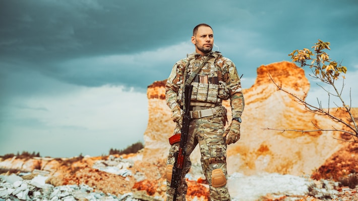 A man in camouflage military gear stands confidently in a rugged, desolate landscape. His attire includes tactical equipment and a rifle slung over his shoulder. The background features dramatic, cloudy skies and rocky, barren terrain with some sparse vegetation.