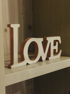 Wooden letters spell out the word 'LOVE' on a shelf. The letters are painted white with a slightly rustic appearance and reddish-brown sides. The shelf is made of light-colored wood, creating a warm and cozy atmosphere.
