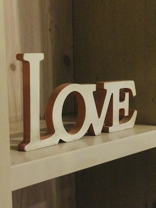 Wooden letters spell out the word 'LOVE' on a shelf. The letters are painted white with a slightly rustic appearance and reddish-brown sides. The shelf is made of light-colored wood, creating a warm and cozy atmosphere.