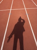 A shadow of a person is cast on a running track marked with white lines. The track surface is a reddish-brown color, and the shadow shows the person with an arm raised near their head.