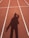 A shadow of a person is cast on a running track marked with white lines. The track surface is a reddish-brown color, and the shadow shows the person with an arm raised near their head.