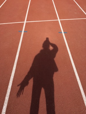A shadow of a person is cast on a running track marked with white lines. The track surface is a reddish-brown color, and the shadow shows the person with an arm raised near their head.
