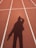 A shadow of a person is cast on a running track marked with white lines. The track surface is a reddish-brown color, and the shadow shows the person with an arm raised near their head.
