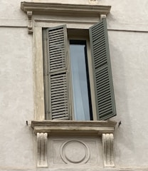 A detailed view of a window set in a stone wall with a partially open wooden shutter. The window is framed by decorative masonry that includes ornate carvings and embellishments. The shutter is painted in a muted green color and casts a shadow on the pale wall.