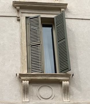A detailed view of a window set in a stone wall with a partially open wooden shutter. The window is framed by decorative masonry that includes ornate carvings and embellishments. The shutter is painted in a muted green color and casts a shadow on the pale wall.