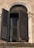 An antique, arched window with intricate architectural detailing, featuring a dark, weathered wooden shutter that is partially open. Behind the shutter, a white curtain is visible, adding a touch of softness to the rustic setting.