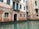 A historic building with a brick facade partially submerged in water. The structure features arched windows with metal bars, and a small balcony with green shutters. The water is calm, reflecting the building's worn texture and muted color palette.