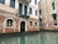 A historic building with a brick facade partially submerged in water. The structure features arched windows with metal bars, and a small balcony with green shutters. The water is calm, reflecting the building's worn texture and muted color palette.