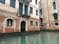 A historic building with a brick facade partially submerged in water. The structure features arched windows with metal bars, and a small balcony with green shutters. The water is calm, reflecting the building's worn texture and muted color palette.