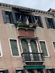 An old building facade with wooden shutters painted in dark green. The upper windows have open shutters with a small balcony decorated with potted plants and a couple of flags. A striped awning partially covers the area above the windows.