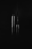 A sleek silver mascara tube stands upright, next to its wand applicator. The lighting emphasizes the metallic texture and reflections on the surface, set against a dark background.