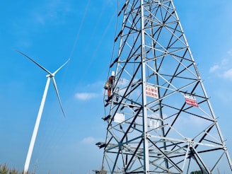 A person is climbing a tall metal electricity tower, which is supported by a complex lattice structure. In the background, there is a large wind turbine with three blades. The sky is clear and blue, suggesting a bright, sunny day. Warning signs are visible on the electricity tower.