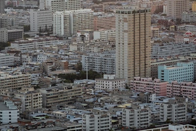 A densely packed urban landscape features numerous residential and commercial buildings of varying heights and designs. Many mid-rise buildings have boxy, geometric shapes, with some showing signs of wear. The tallest structure is a prominent high-rise building located near the center. The scene conveys a sense of bustling city life and urban development.