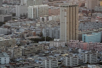 A densely packed urban landscape features numerous residential and commercial buildings of varying heights and designs. Many mid-rise buildings have boxy, geometric shapes, with some showing signs of wear. The tallest structure is a prominent high-rise building located near the center. The scene conveys a sense of bustling city life and urban development.