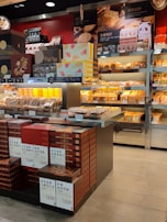 A retail display features a variety of packaged baked goods, including mooncakes and other traditional pastries. The packages are neatly stacked on shelves and tables, with some boxes showcasing intricate designs. Signage in a language possibly indicating product names and prices is visible. The display is well-lit, highlighting the vibrant packaging and colorful product boxes.
