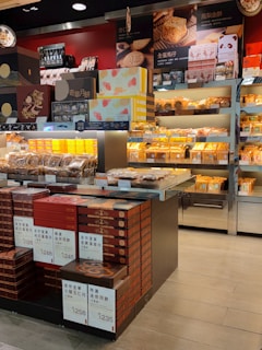 A retail display features a variety of packaged baked goods, including mooncakes and other traditional pastries. The packages are neatly stacked on shelves and tables, with some boxes showcasing intricate designs. Signage in a language possibly indicating product names and prices is visible. The display is well-lit, highlighting the vibrant packaging and colorful product boxes.