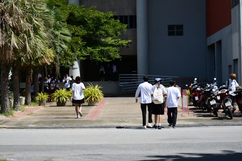 A group of people wearing white shirts are walking towards a building with large columns. There are motorcycles parked on the right side, and lush green trees and bushes on the left. The atmosphere suggests a school or university environment.