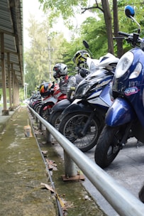 A row of parked motorcycles, some with helmets attached to them, positioned under an outdoor shelter with a metal roof. The motorcycles are lined up closely along a metal railing, with a green, leafy backdrop and power lines visible in the distance. The ground near the motorcycles is concrete, and there are leaves scattered around.