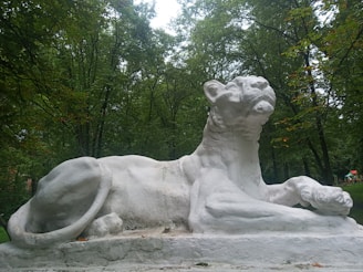 A large, white stone sculpture of a reclining lion is situated in a lush, green forested area. The lion's pose is majestic, with its head raised and paws stretched out in front. Surrounding the sculpture are numerous tall trees with dense foliage.