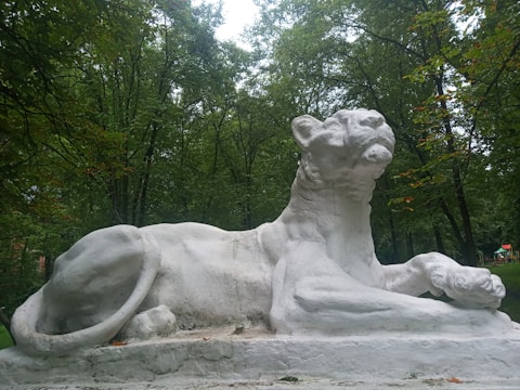 A large, white stone sculpture of a reclining lion is situated in a lush, green forested area. The lion's pose is majestic, with its head raised and paws stretched out in front. Surrounding the sculpture are numerous tall trees with dense foliage.