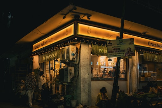 A dimly lit street corner scene featuring a shop called 'Green Neko' with a glowing yellow sign. Two people are standing near the entrance, and the shop has visible signs for 'BOBA' and 'SIBA TEA SPOKEBOJ'. Air conditioning units are mounted outside the shop. A poster with images and text in an unknown language is attached to a pole, and there are some plants near the entrance. The atmosphere suggests an evening or nighttime setting.