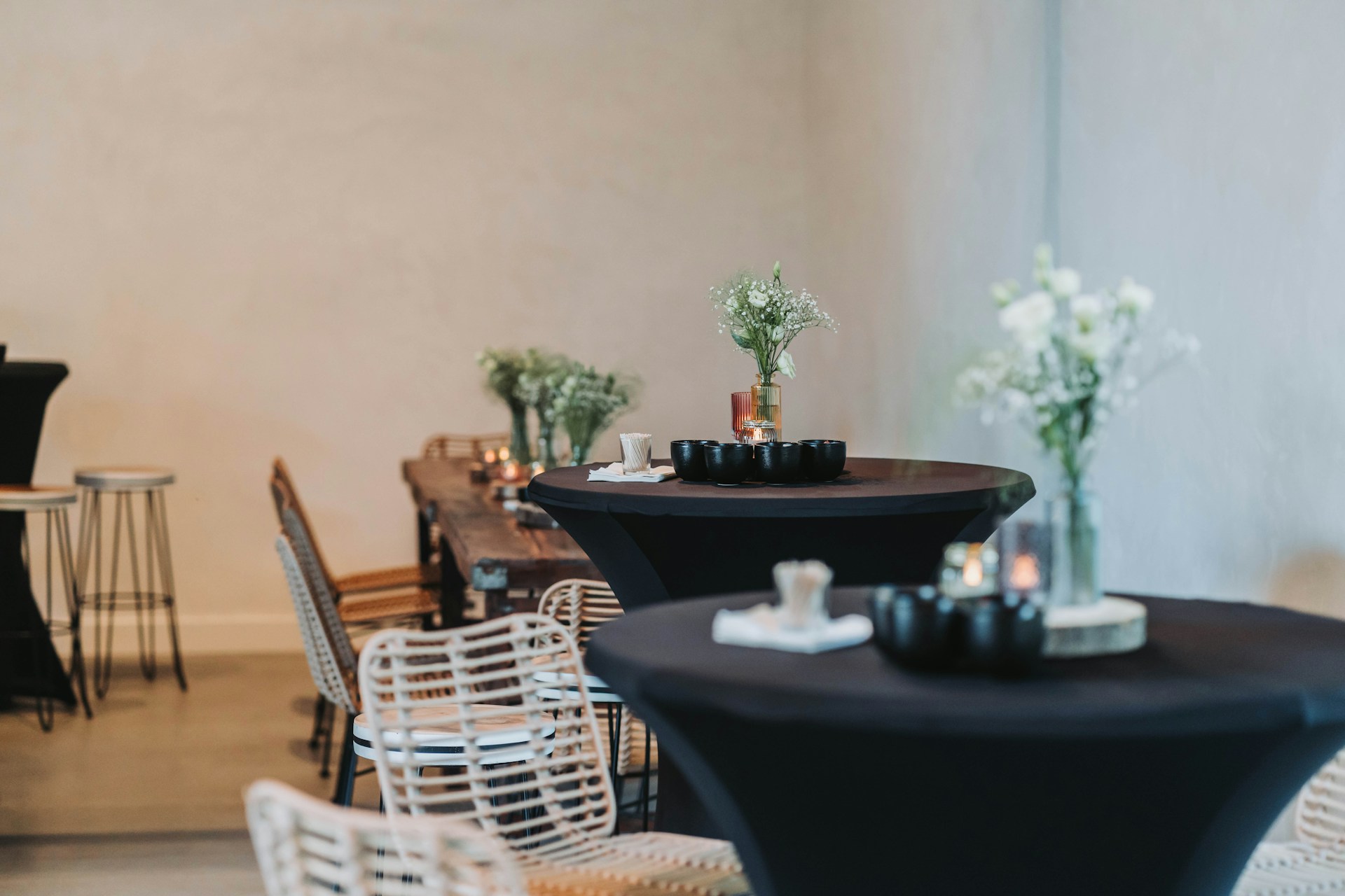 A dining area with dark tables set up with small black bowls, candles, and jars of white flowers. There are white wicker chairs around the tables, and a long wooden communal table with similar decor. The atmosphere is minimalist and elegant, with neutral walls and soft lighting.